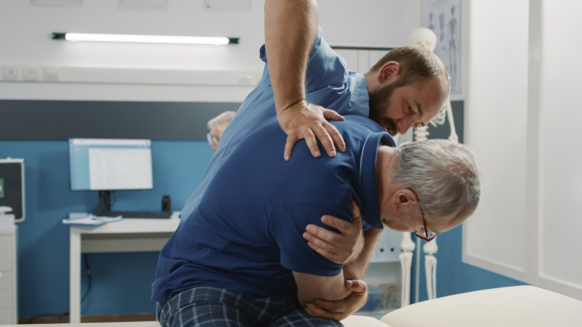 Osteopath helping senior man to crack back bones in cabinet, doing physical therapy exercise. Male assistant using physiotherapy procedure to increase mobility and treat patient.